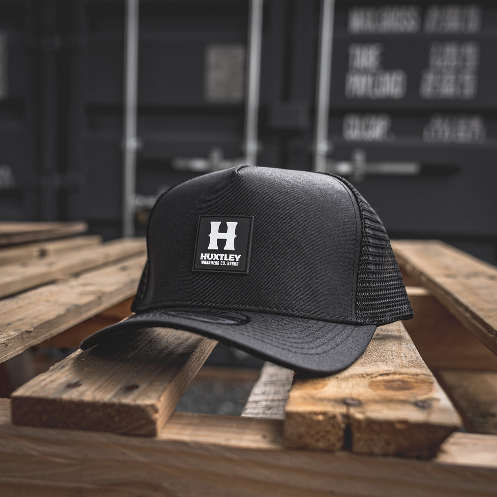 Close-up of a black Huxtley Workwear trucker cap featuring a bold rubber 'H' logo patch—built for durability and designed for hard wear on the job or off-duty.
