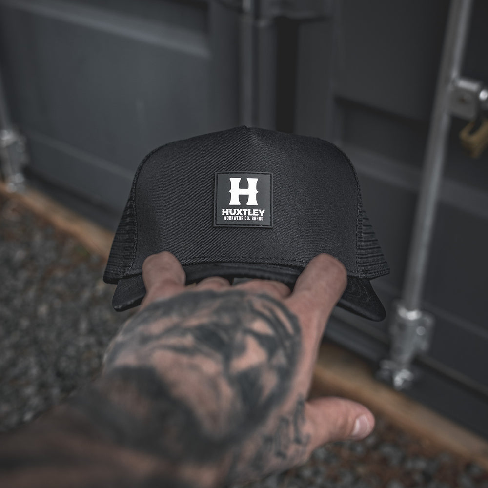 
                  
                    Front view of the Huxtley trucker cap featuring a bold rubber patch logo. Minimalist and durable, this structured cap combines streetwear edge with work-ready reliability.
                  
                
