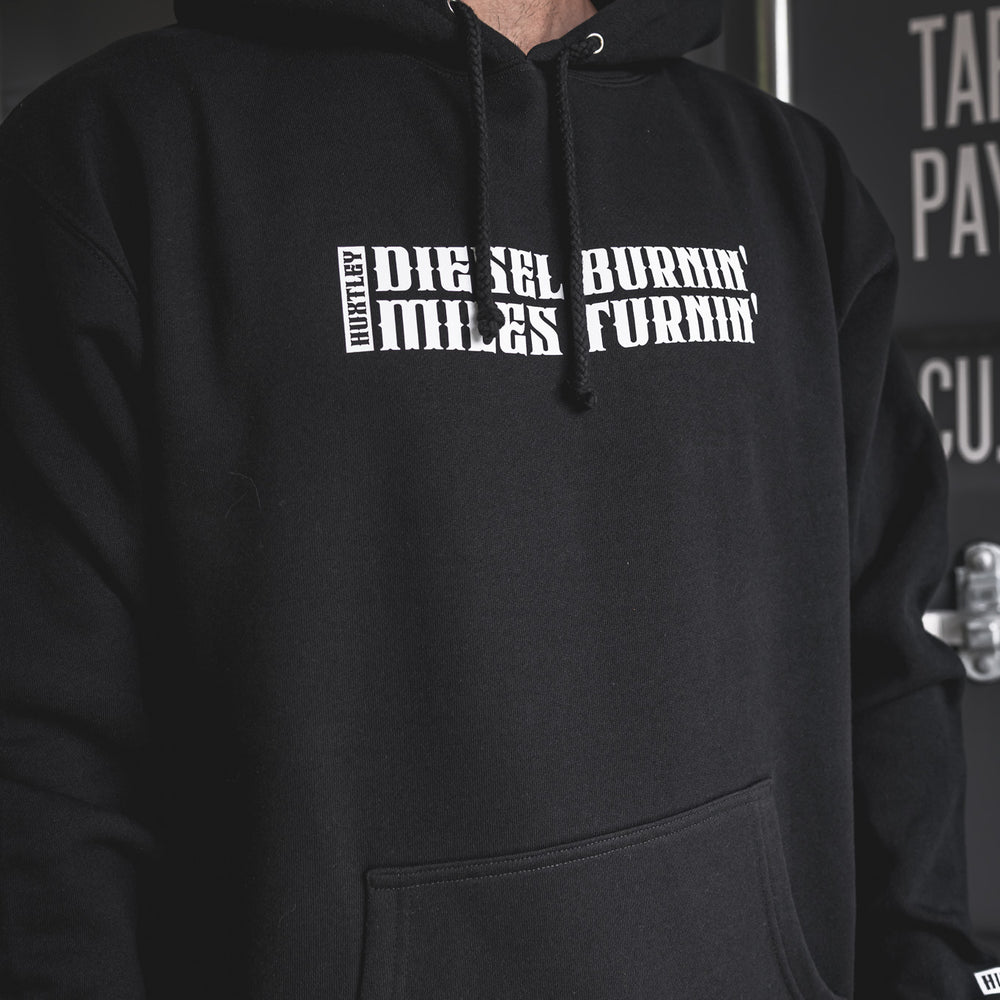
                  
                    Close-up of black hoodie with “Diesel Burnin’ Miles Turnin’” chest graphic and woven Huxtley tag on the cuff. Workwear fit with bold attitude.
                  
                
