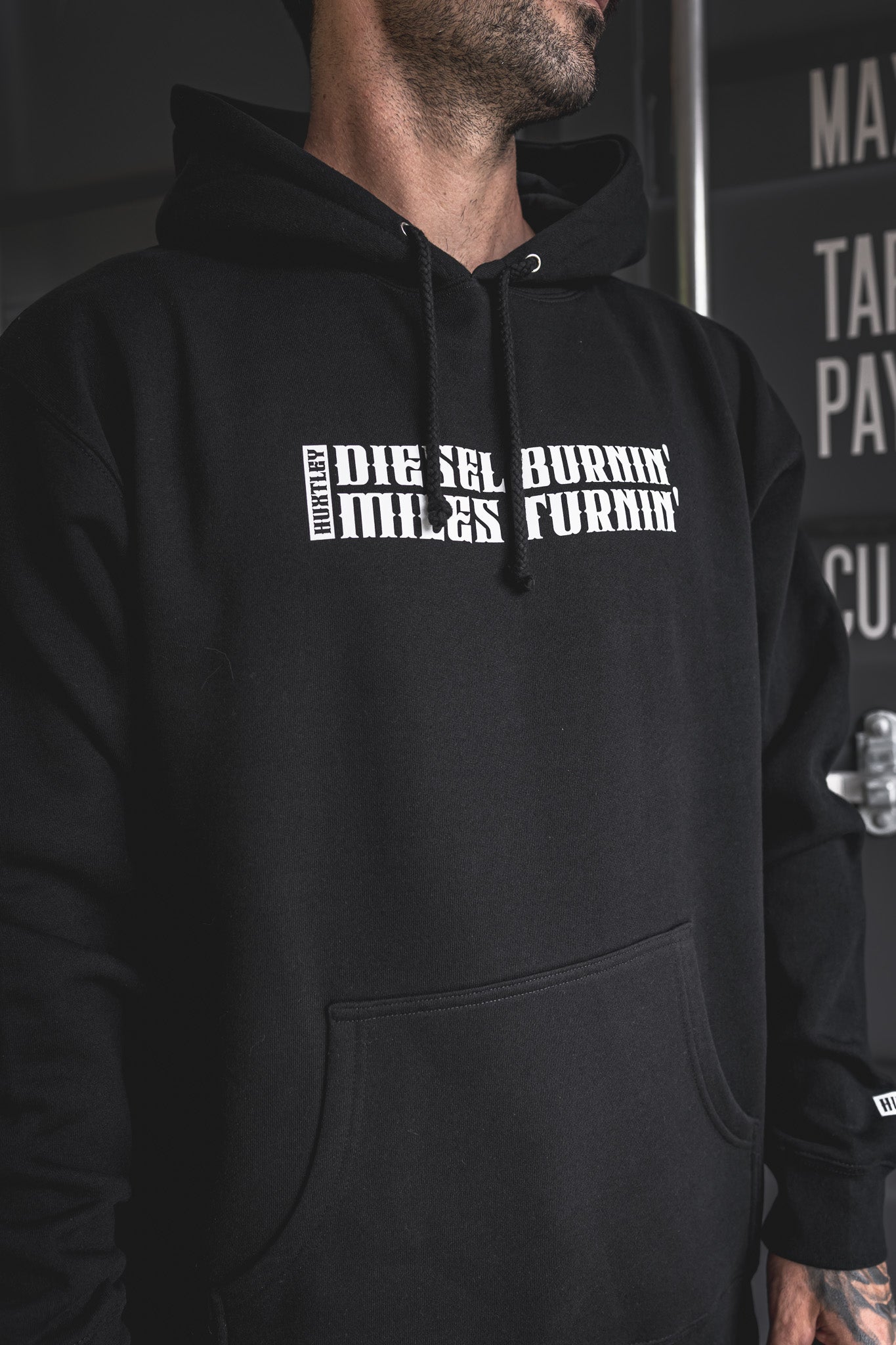 
                  
                    Close-up of black hoodie with “Diesel Burnin’ Miles Turnin’” chest graphic and woven Huxtley tag on the cuff. Workwear fit with bold attitude.
                  
                