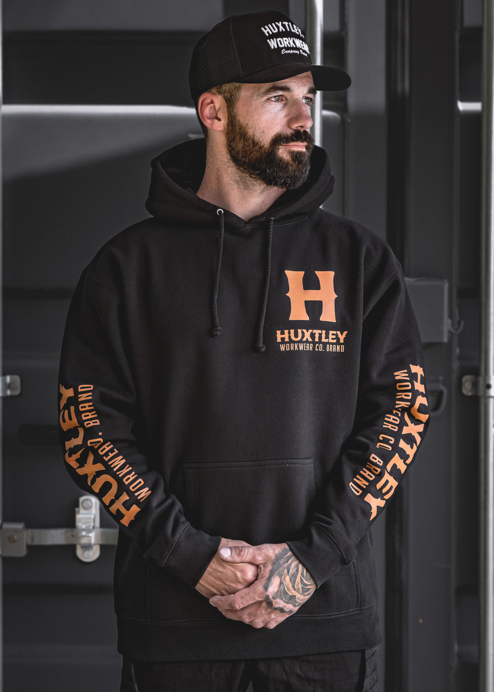 The Full Print Hoodie by Huxtley Workwear features a bold orange logo on the chest and both sleeves. Shown worn in an industrial setting, this black pullover blends comfort, durability, and standout style for the jobsite or the street.