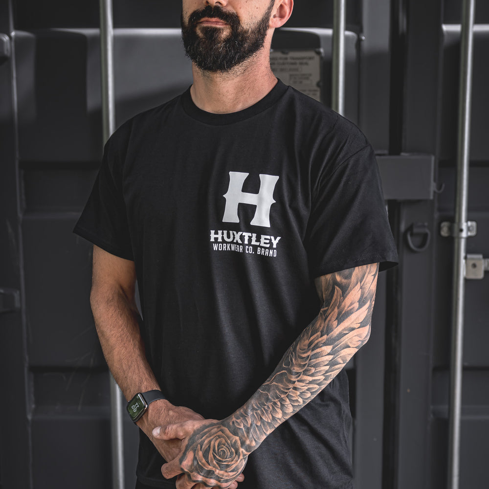 
                  
                    Model wearing the Huxtley Full Print tee in classic black, featuring a bold white front logo. Built for grit and clean looks.
                  
                