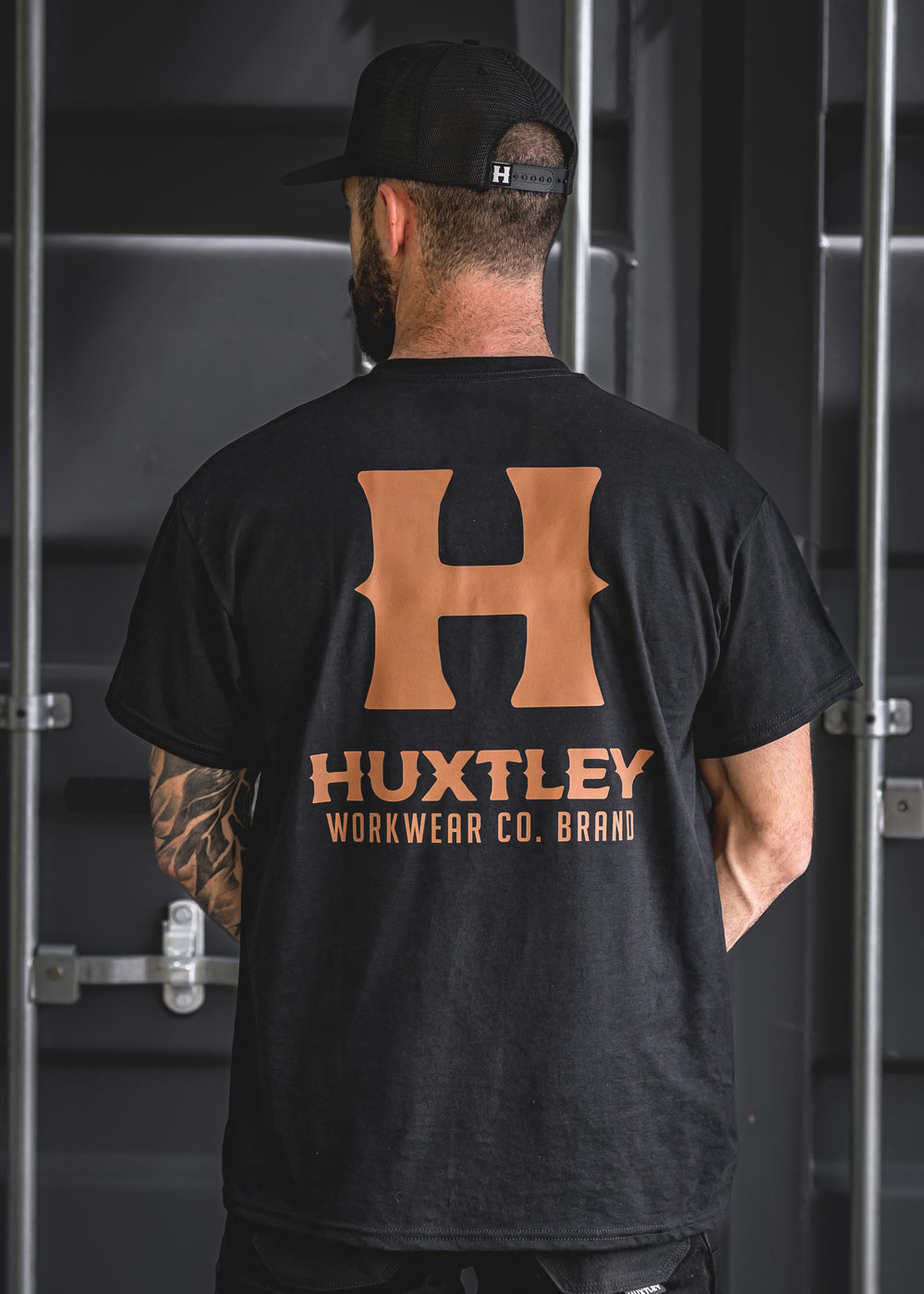 Back view of the Huxtley Full Print tee with oversized whiskey-colored “H” and logo. Bold workwear branding on classic black cotton.