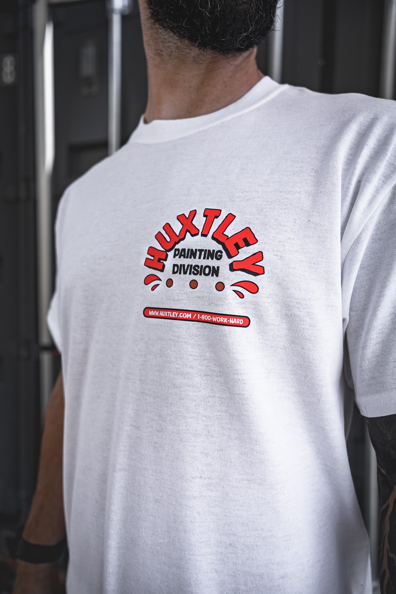 
                  
                    Close-up of the Huxtley Painting Division graphic on a crisp white work tee. Bold red and black print stamped across the chest, complete with the hotline and website for the real ones who paint and hustle hard.
                  
                