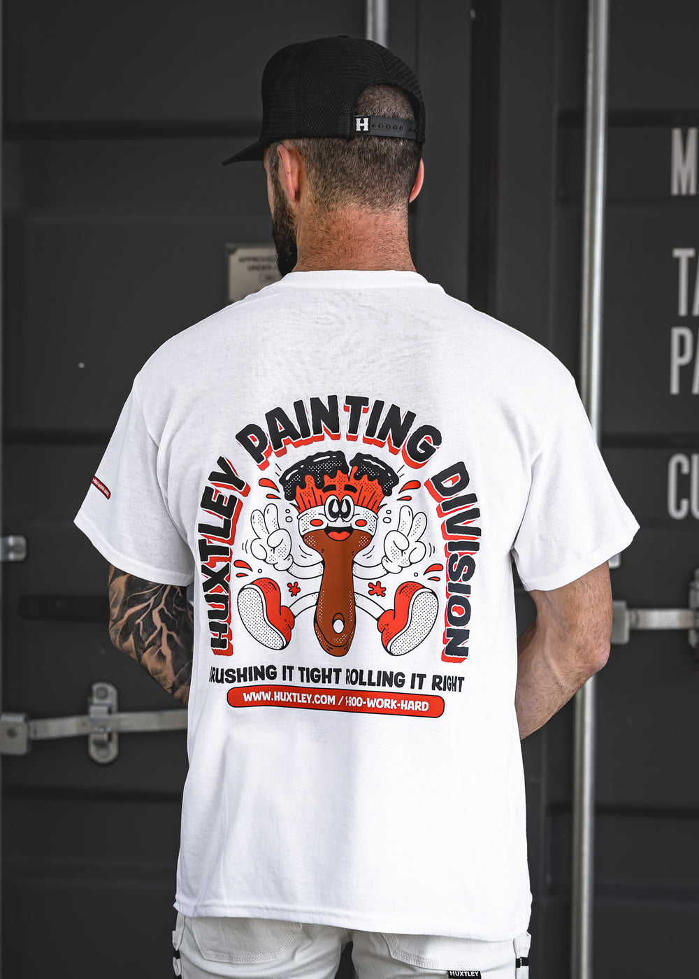White t-shirt from the Huxtley Painting Division, featuring a bold back graphic of a cartoon brush and slogan: “Brushing it tight, rolling it right.” Perfect for painters who take pride in the craft.