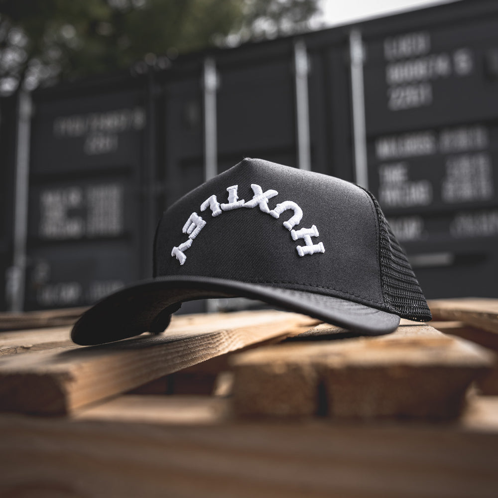 
                  
                    Front-focused view of the Huxtley Reverse Logo cap. Signature arched embroidery pops against the classic black, laid out on rugged pallet wood with shipping containers in the background.
                  
                