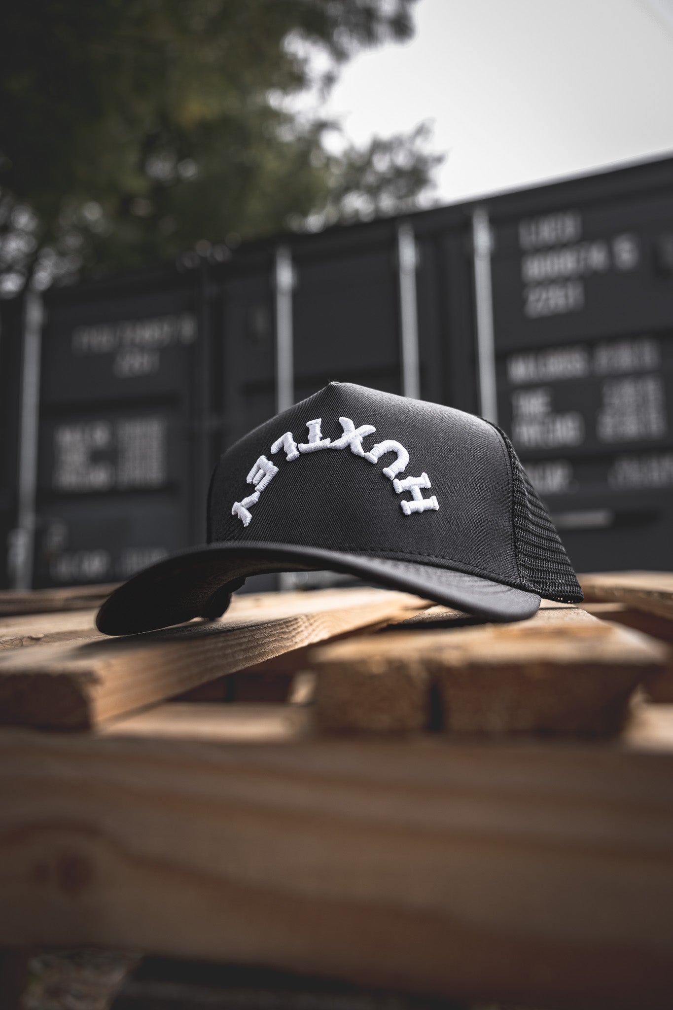 
                  
                    Front-focused view of the Huxtley Reverse Logo cap. Signature arched embroidery pops against the classic black, laid out on rugged pallet wood with shipping containers in the background.
                  
                