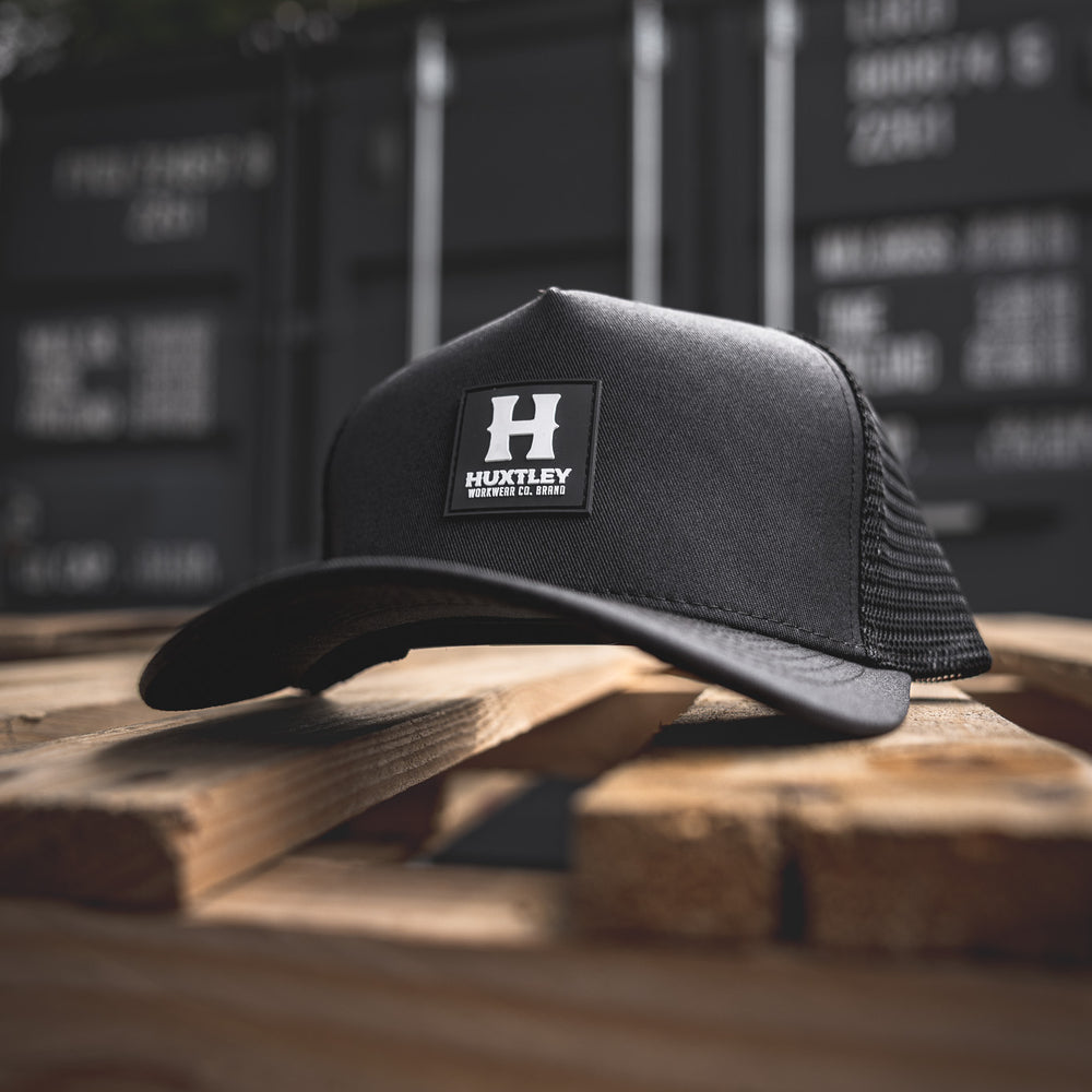 
                  
                    Angled view of the Huxtley trucker cap resting on a rugged wood pallet. Features a bold rubber patch logo and mesh back, built tough for the jobsite and beyond.
                  
                