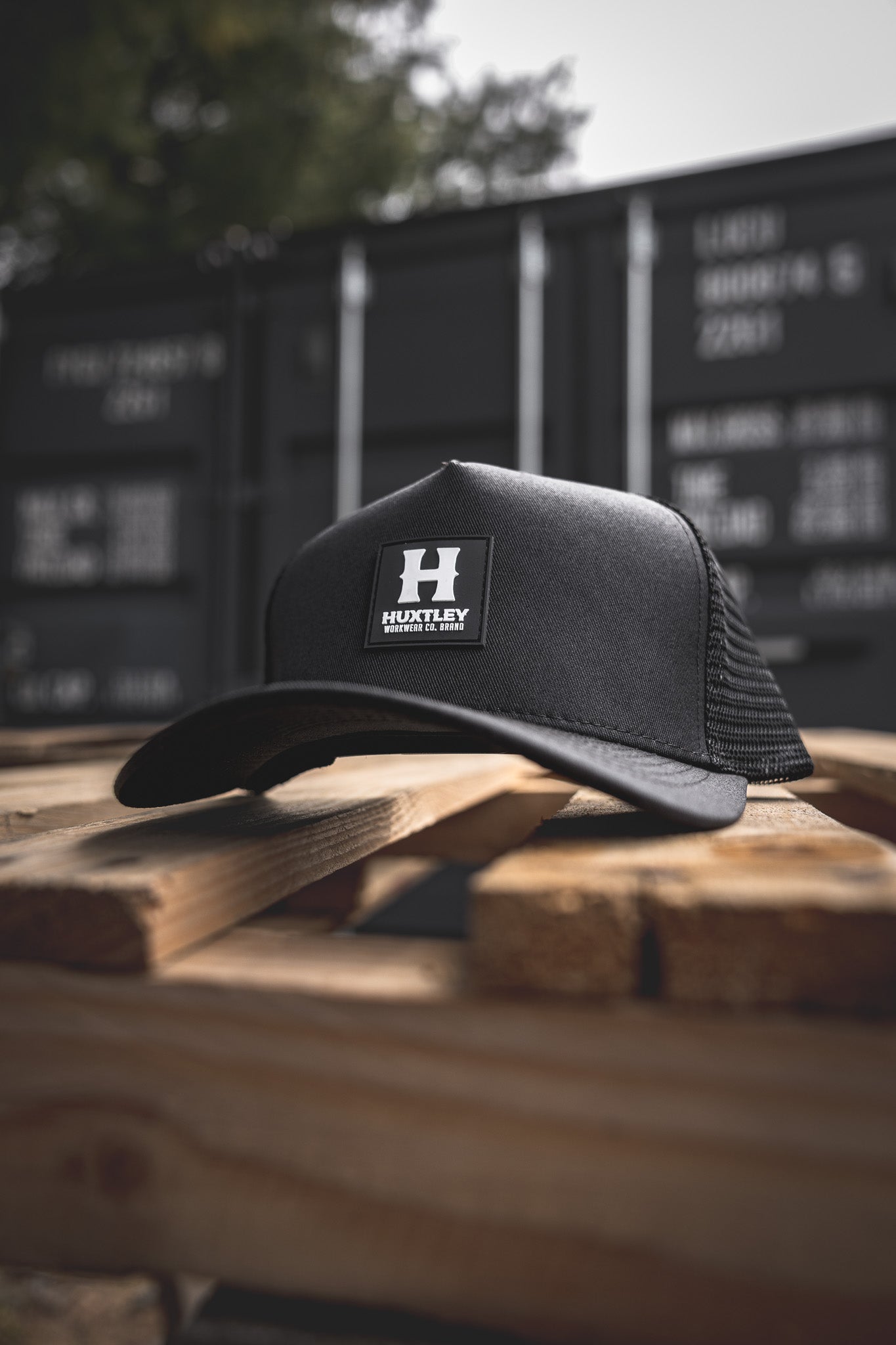 
                  
                    Angled view of the Huxtley trucker cap resting on a rugged wood pallet. Features a bold rubber patch logo and mesh back, built tough for the jobsite and beyond.
                  
                