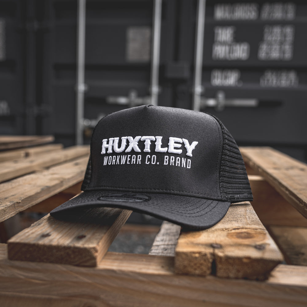 Close-up of the Huxtley Workwear Co. Brand cap resting on a rugged wood pallet. Bold white embroidery stands out on a structured black trucker frame, built for tough days on-site.