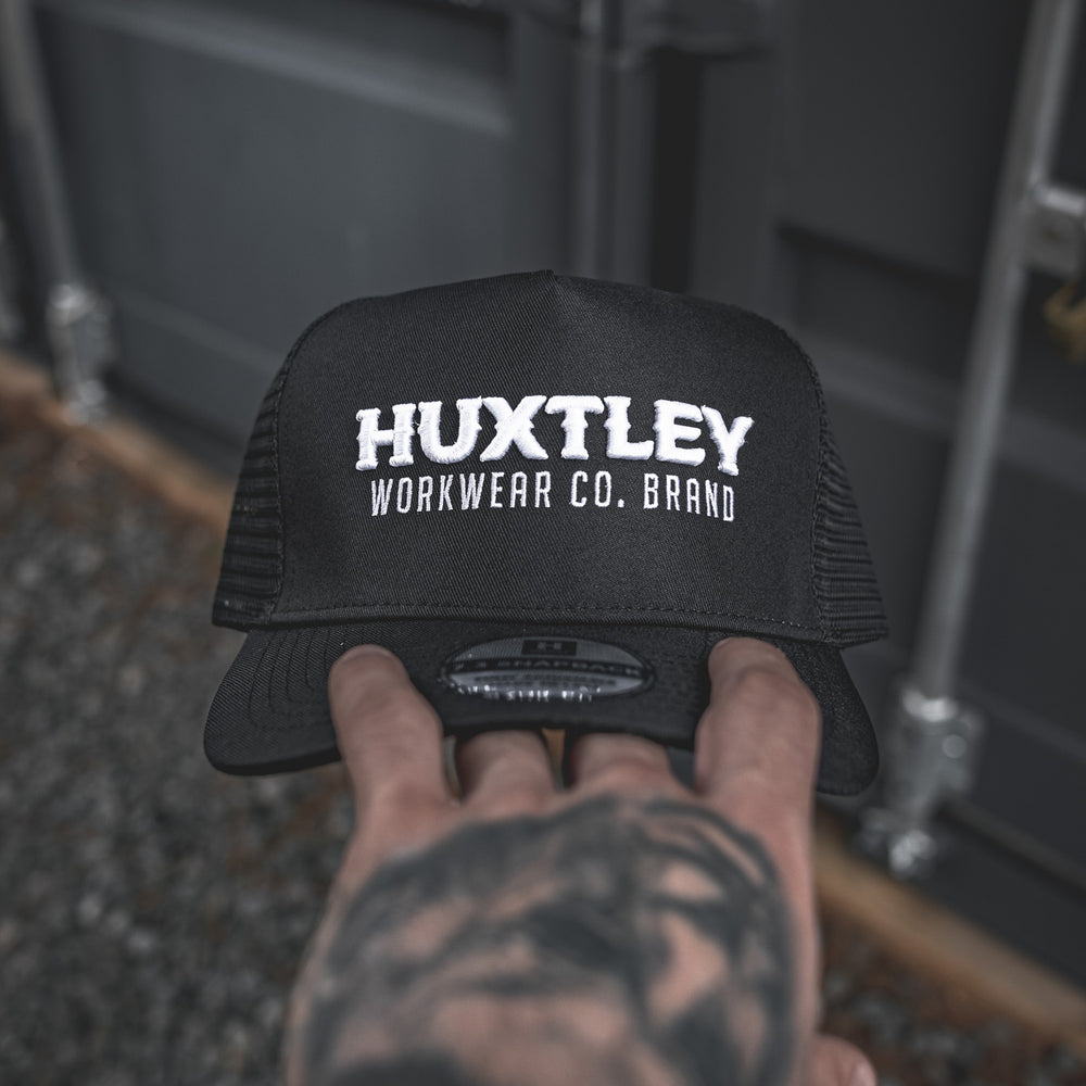 
                  
                    Close-up of the Huxtley Workwear Co. Brand trucker cap front logo. Embroidered in bold white on black, this cap means business—on site and off.
                  
                