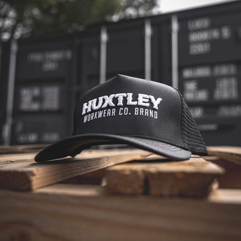 
                  
                    The Huxtley Workwear Co. Brand trucker cap sits front and center on a rugged pallet, with shipping containers in the background—built for tough days on site.
                  
                