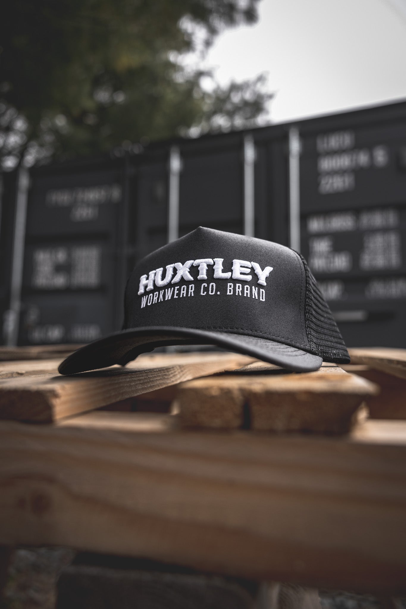 
                  
                    The Huxtley Workwear Co. Brand trucker cap sits front and center on a rugged pallet, with shipping containers in the background—built for tough days on site.
                  
                