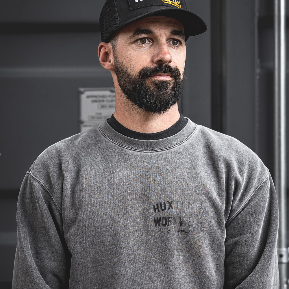 
                  
                    Model wearing the black mesh Huxtley trucker cap featuring the bold "Work Hard" patch. Paired with a washed-out crewneck in front of a steel container, this look embodies jobsite grit and blue-collar pride.
                  
                