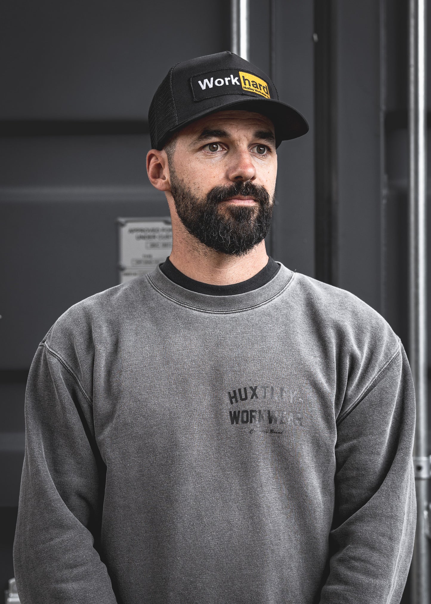 
                  
                    Model wearing the black mesh Huxtley trucker cap featuring the bold "Work Hard" patch. Paired with a washed-out crewneck in front of a steel container, this look embodies jobsite grit and blue-collar pride.
                  
                