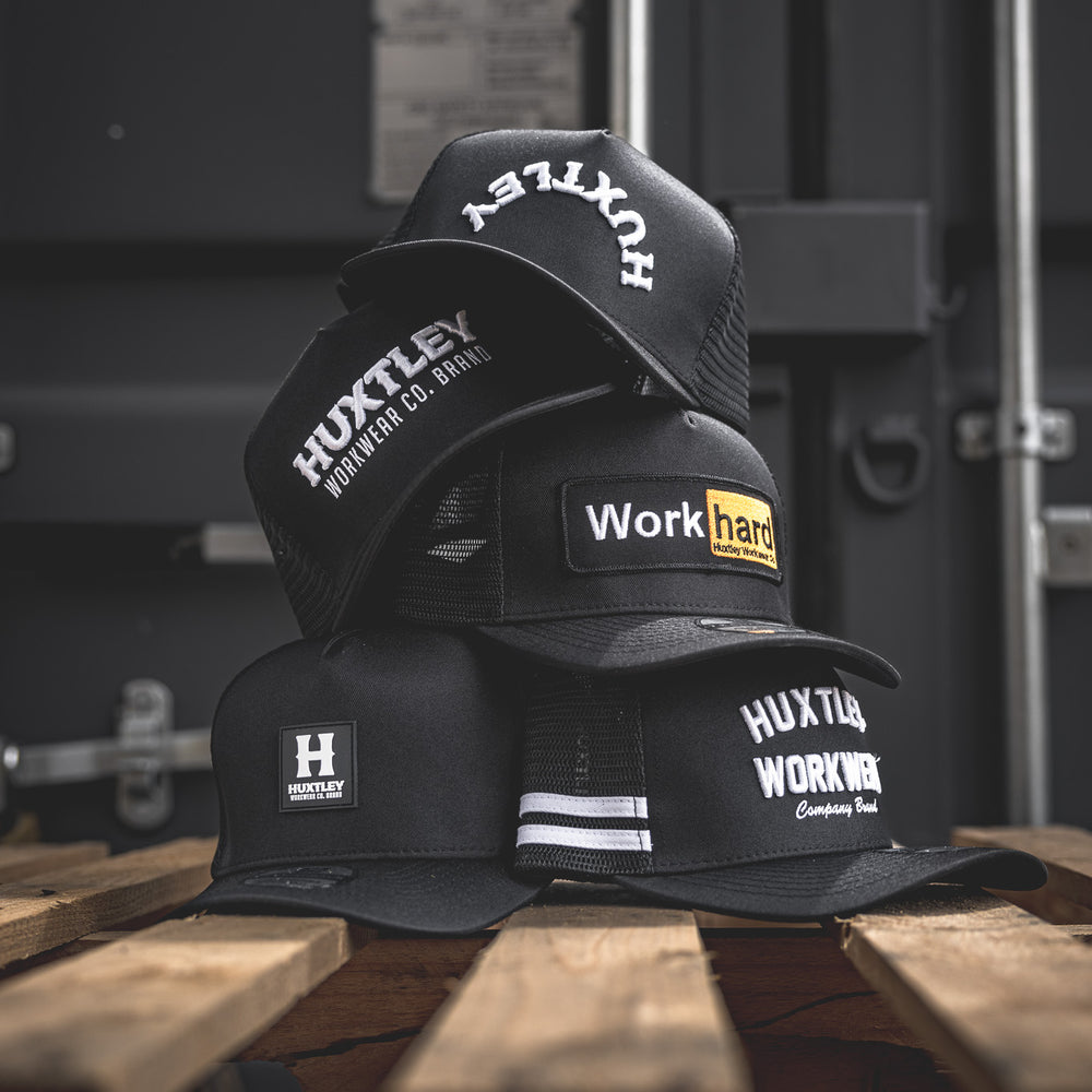 
                  
                    A bold lineup of Huxtley Workwear trucker caps stacked on a rugged pallet—featuring multiple embroidered designs that speak grit, grind, and on-site pride.
                  
                