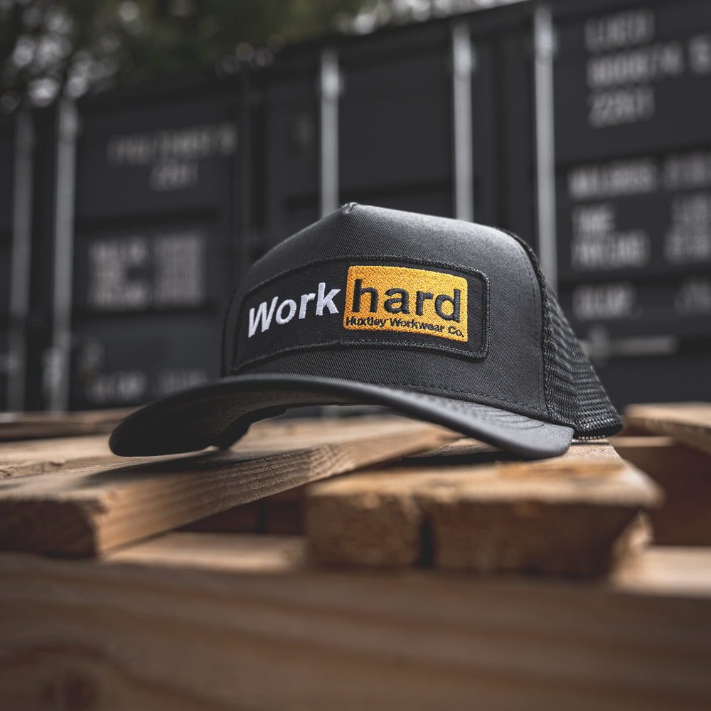 
                  
                    Close-up of the Huxtley “Work hard” mesh-back cap, shot outdoors on a rustic pallet with black shipping containers in the background. Rugged, real, ready to hustle.
                  
                