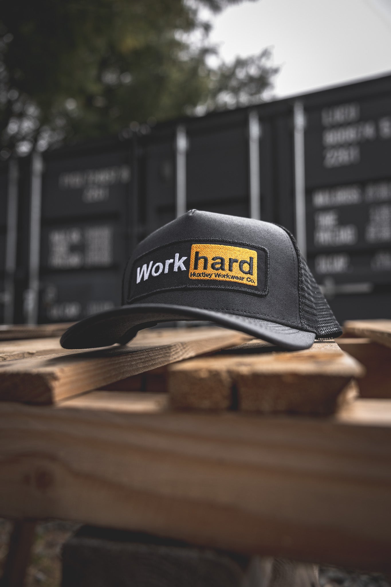 
                  
                    Close-up of the Huxtley “Work hard” mesh-back cap, shot outdoors on a rustic pallet with black shipping containers in the background. Rugged, real, ready to hustle.
                  
                